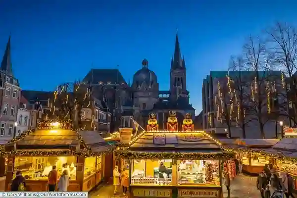 Marché de Noël d’Aachen. Elle montre les étals du marché installés devant la cathédrale d’Aachen. L’image capture magnifiquement l’atmosphère festive avec l’éclairage chaud des étals contrastant avec le ciel crépusculaire.