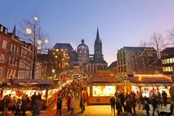 Marché de Noël d'Aix-la-Chapelle (Aachen) en Allemagne, illuminé le soir. L'image capture bien l'atmosphère chaleureuse et festive du marché, avec les nombreuses cabanes en bois, les lumières scintillantes et la cathédrale d'Aix-la-Chapelle en arrière-plan. On voit également de nombreuses personnes se promenant et profitant de l'ambiance.