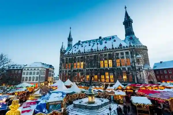 Marché de Noël d’Aix-la-Chapelle (Aachener Weihnachtsmarkt) sur le Markt (place du marché). Le Rathaus (hôtel de ville) est le bâtiment dominant à l’arrière-plan. L’image met en valeur l’atmosphère festive avec les nombreux étals du marché et la scène enneigée.